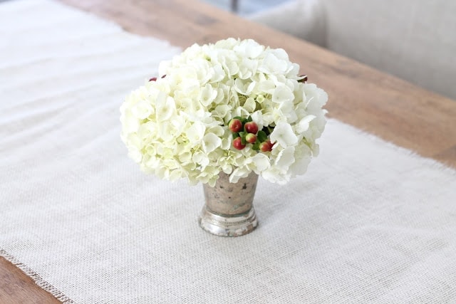 A far shot of white flowers with berry accents in a small vase on a wooden table.