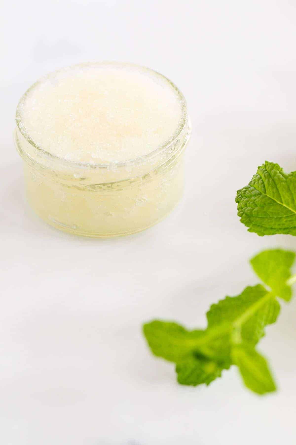 A glass jar full of a sugar scrub recipe, stem of fresh mint nearby.