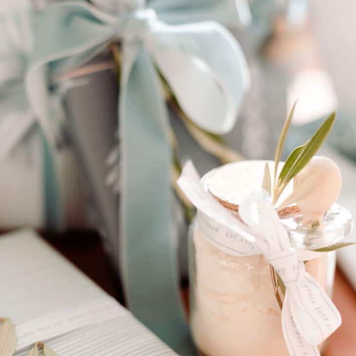 Soft blue and white gift wrapping under a tree, with a clear glass jar of bath salts tied with a bow.
