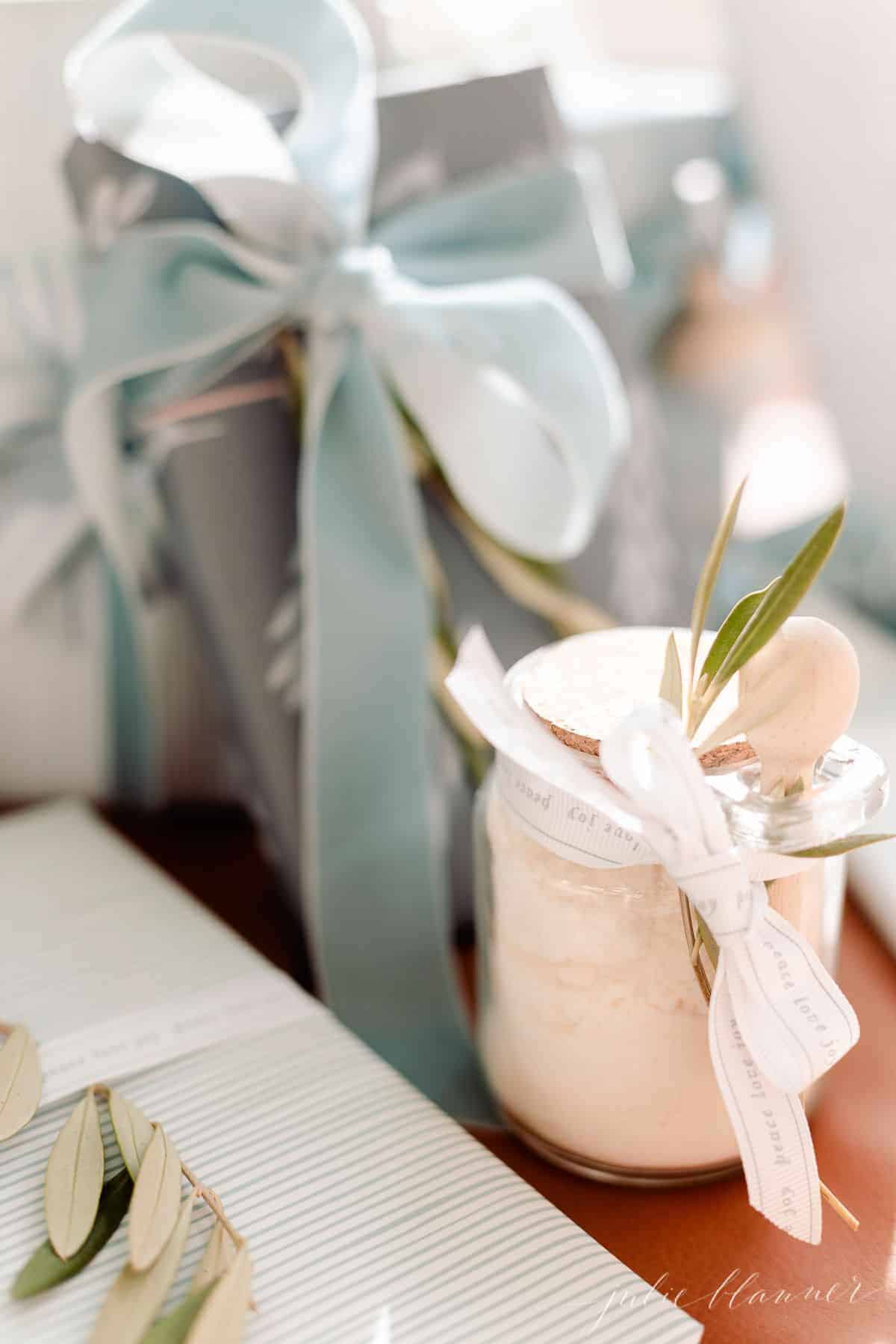 Soft blue and white gift wrapping under a tree, with a clear glass jar of bath salts tied with a bow.