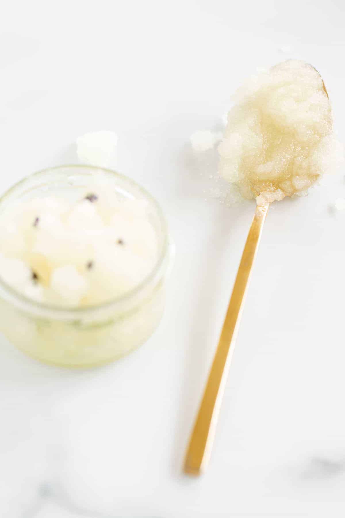 A homemade sugar scrub with lavender, in a glass jar on a white surface, gold spoon to the side.
