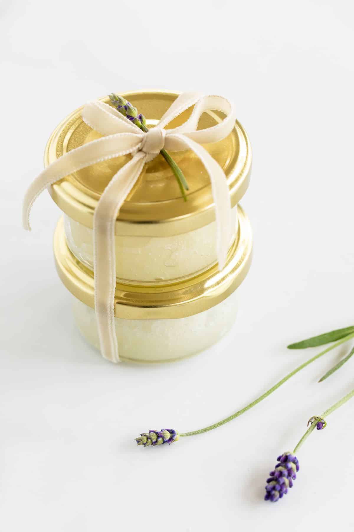 Two small glass bottles with gold lids full of sugar scrub, tied with a bow, fresh lavender in foreground.