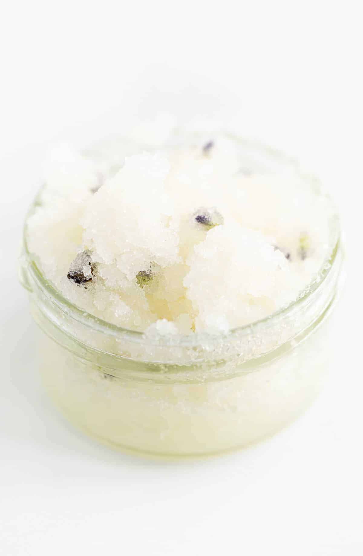 A homemade sugar scrub with lavender, in a glass jar on a white surface.