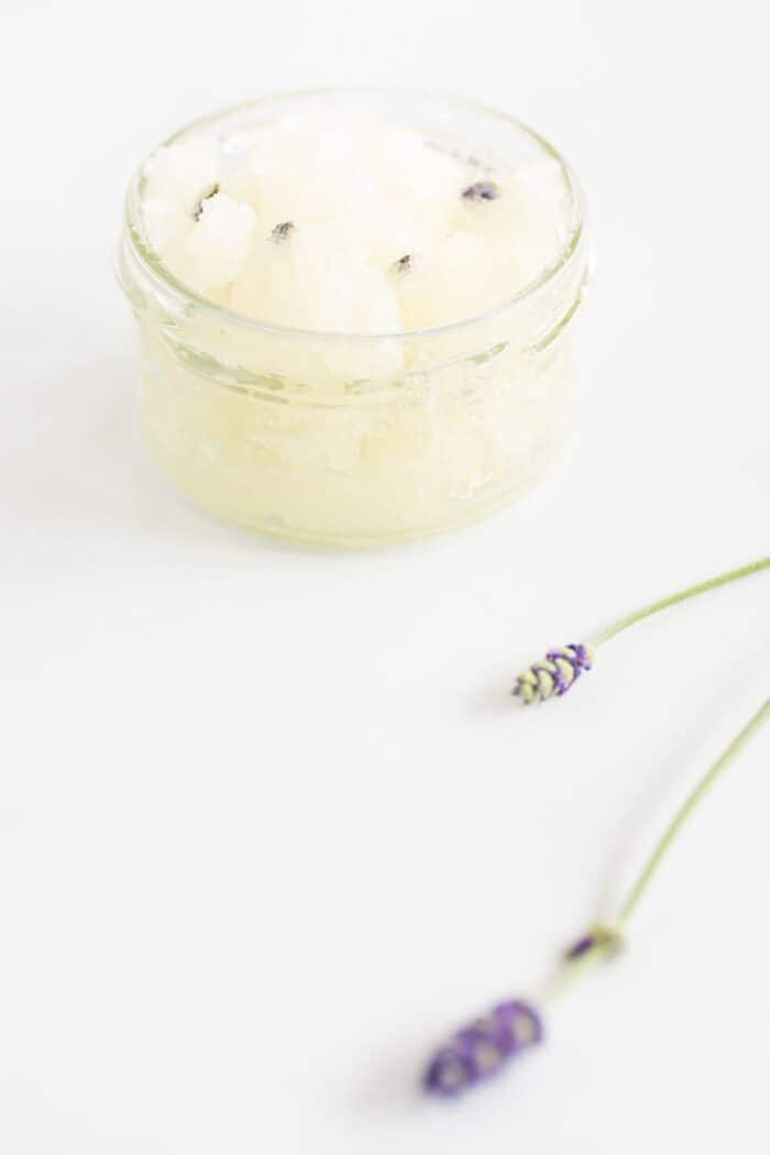 A homemade sugar scrub with lavender, in a glass jar on a white surface.
