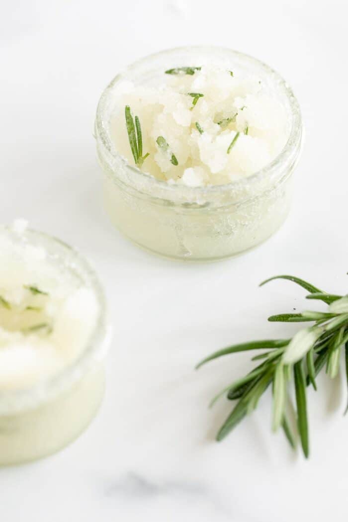 Two jars of homemade sugar scrub on a marble surface, stem of fresh rosemary in foreground.