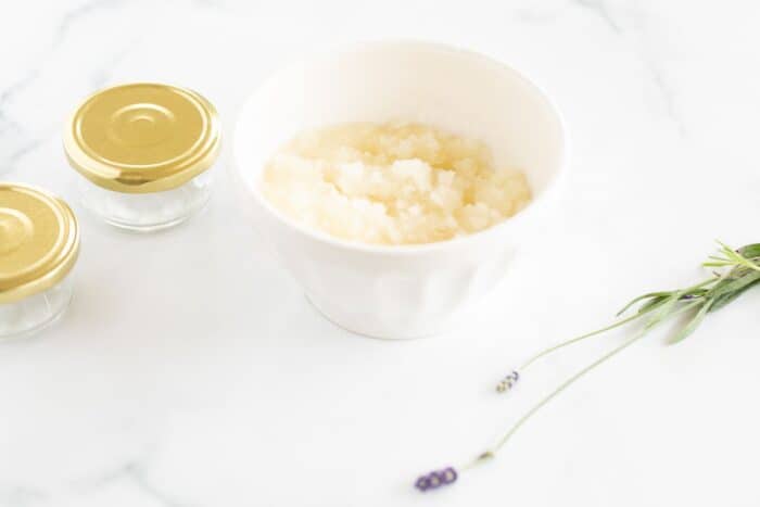 DIY Sugar Scrub made in your kitchen! Great gift idea via www.julieblanner.com Bowl of sugar and clear glass bottles set out on a white surface, fresh lavender stem in foreground.