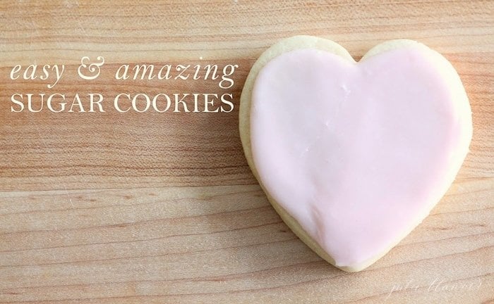 An easy sugar cookie in the shape of a heart with pink frosting, on a wooden surface. Easy & Amazing sugar cookies in a white text overlay.