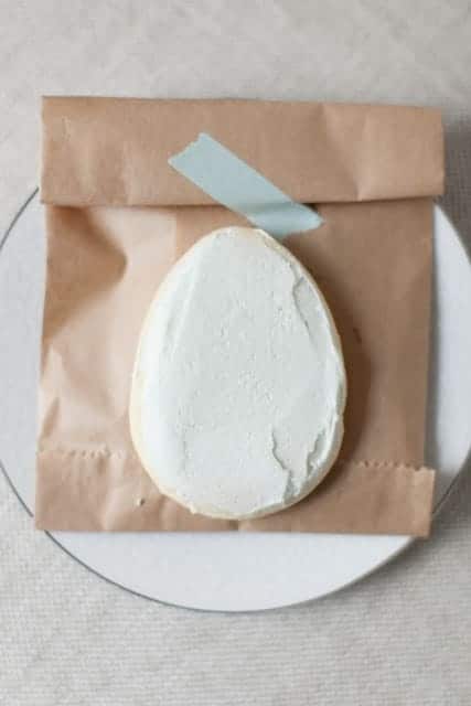 A white frosted sugar cookie in the shape of an egg, on a brown paper treat bag.