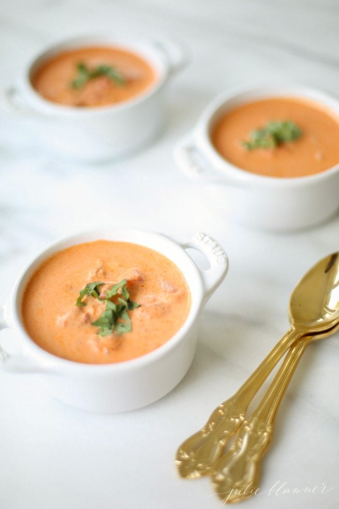 A bowl of tomato soup, with more bowls in the background and a gold spoon to the side.