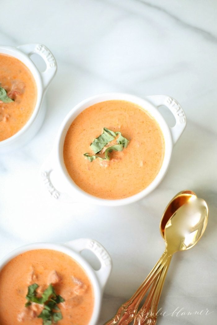 three white coccotes with homemade tomato soup garnished with fresh basil and gold spoons