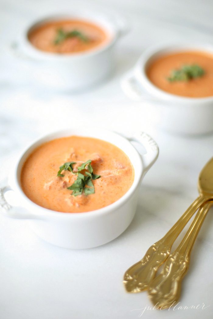 three white coccotes with homemade tomato soup garnished with fresh basil and gold spoons