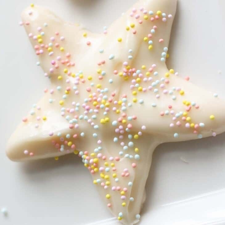 A white star shaped sugar cookie on a white surface, topped with sugar cookie icing and pastel sprinkles.