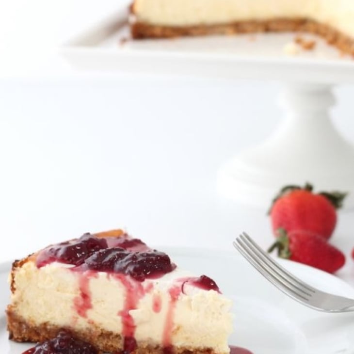 A slice of New York cheesecake on a white plate, with the whole cheesecake on a white cake pedestal in the background.