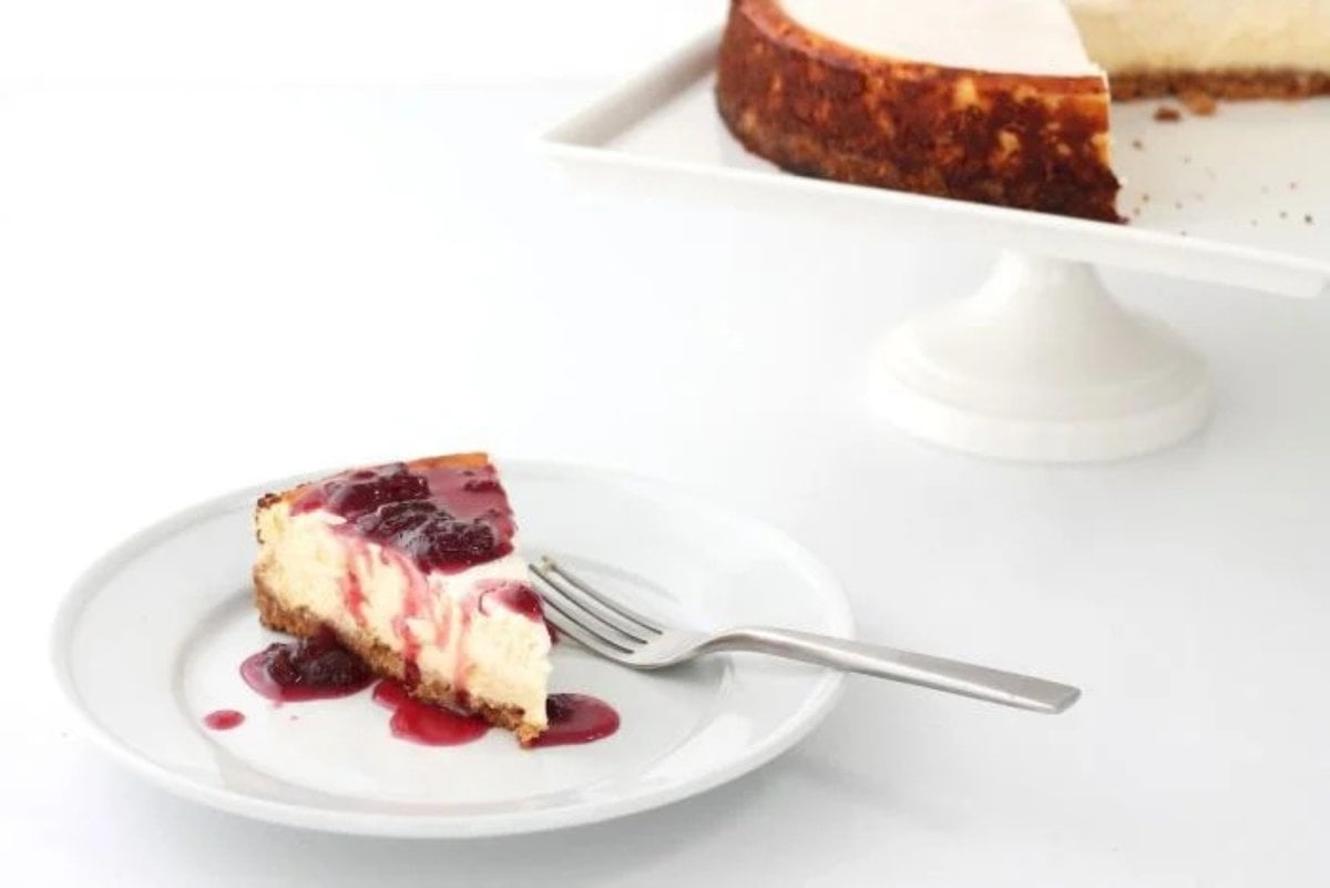 A slice of New York cheesecake on a white plate, with the whole cheesecake on a white cake pedestal in the background.