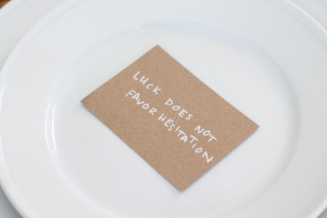A craft paper card on top of a white place setting, handwritten in white ink, reads, "Luck does not favor hesitation."