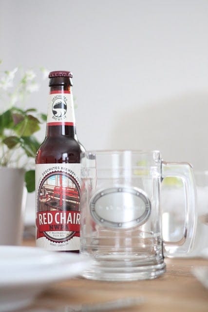 Bottle of Red Chair beer next to a glass mug with handle, sitting on a wooden tabletop with white dishes and clover plant. 