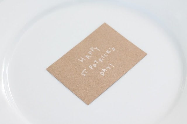 Small handmade craft paper card that reads "Happy St. Patrick's Day!" sits on top of a white plate.
