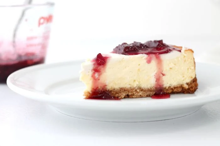 A slice of New York cheesecake on a white plate, with a glass container of strawberry topping in the background.