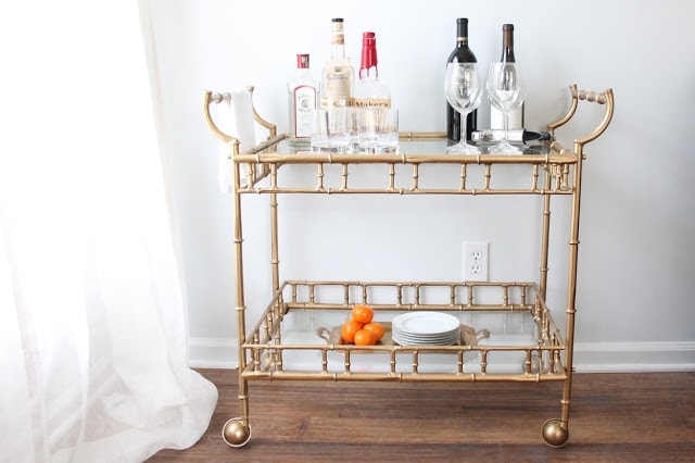 A vintage brass bar cart with glass shelves, topped with liquor bottles and glassware.