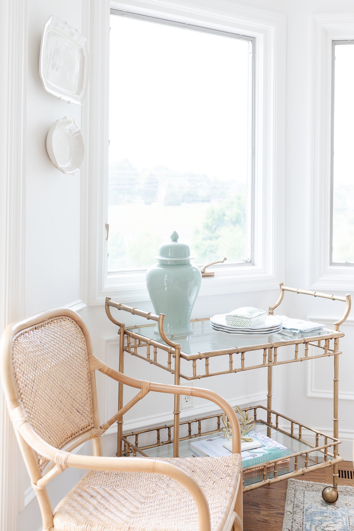 A gold bar cart in a white room with a rattan dining chair next to it.