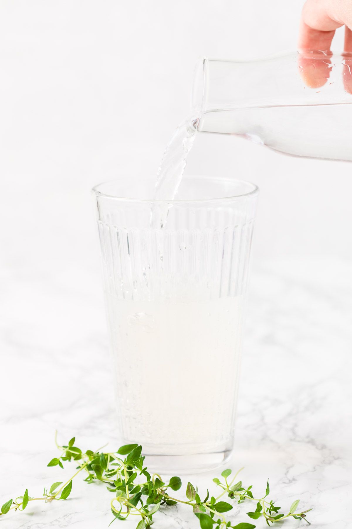 A vodka lemonade cocktail being poured into a clear glass, fresh thyme on the countertop in front
