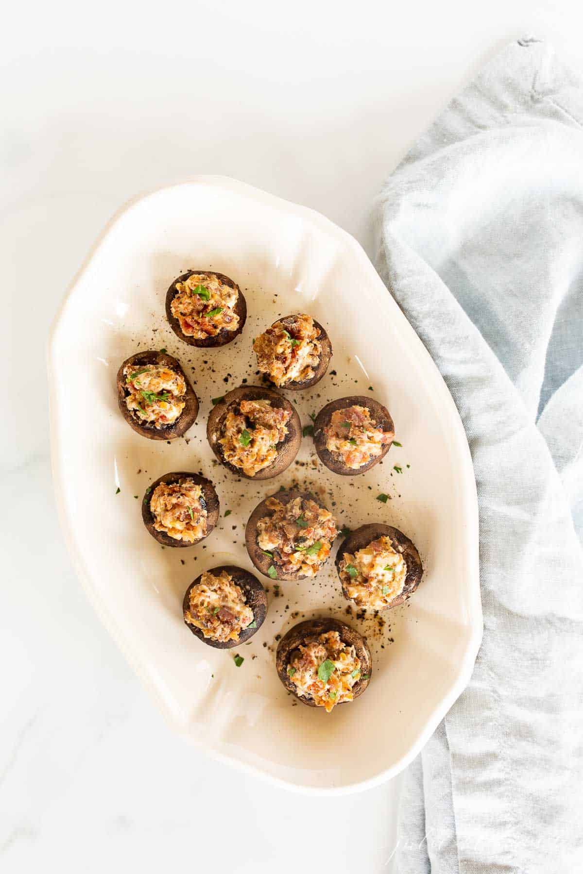 A white oval platter full of cream cheese stuffed mushrooms, blue linen towel to the side.