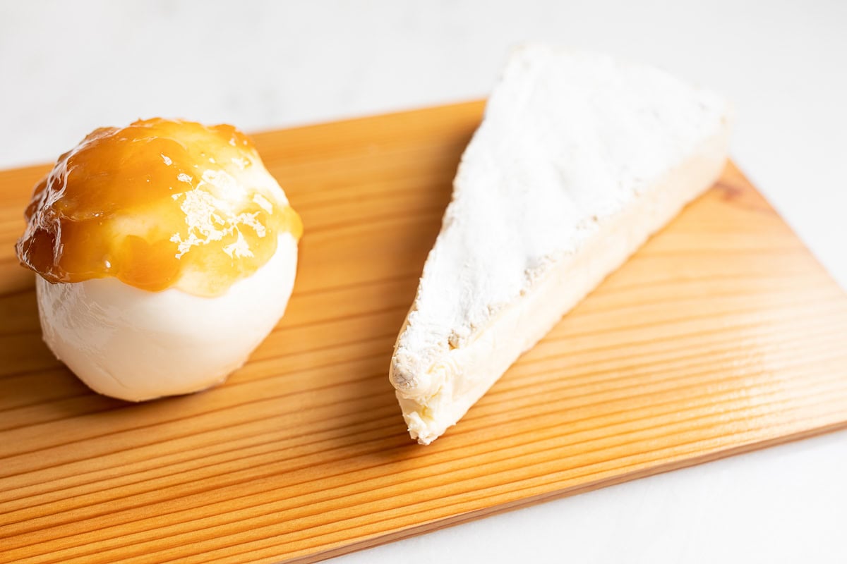 A burrata cheese topped with apricot jam and a wedge of brie cheese, served as a grilled Mozzarella appetizer on a wooden cutting board.
