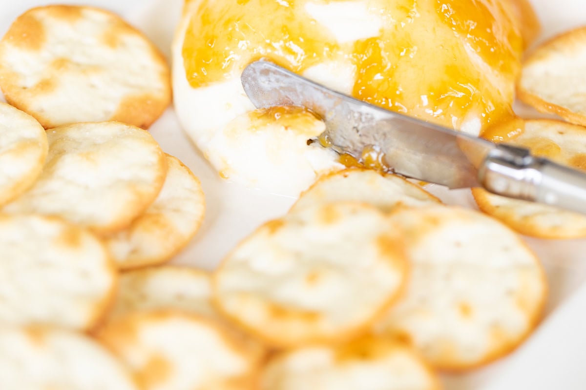 A close-up of a grilled mozzarella cheese ball topped with apricot jam, served with a spreader knife and surrounded by round crackers.