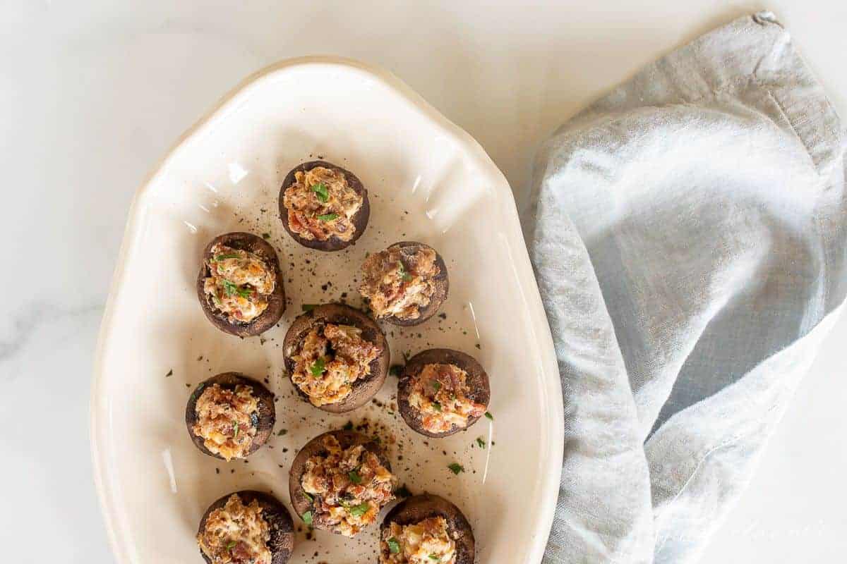 A white oval platter full of cream cheese stuffed mushrooms, blue linen towel to the side.