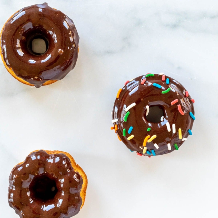 Baked donuts topped with chocolate frosting and sprinkles, on a marble countertop.