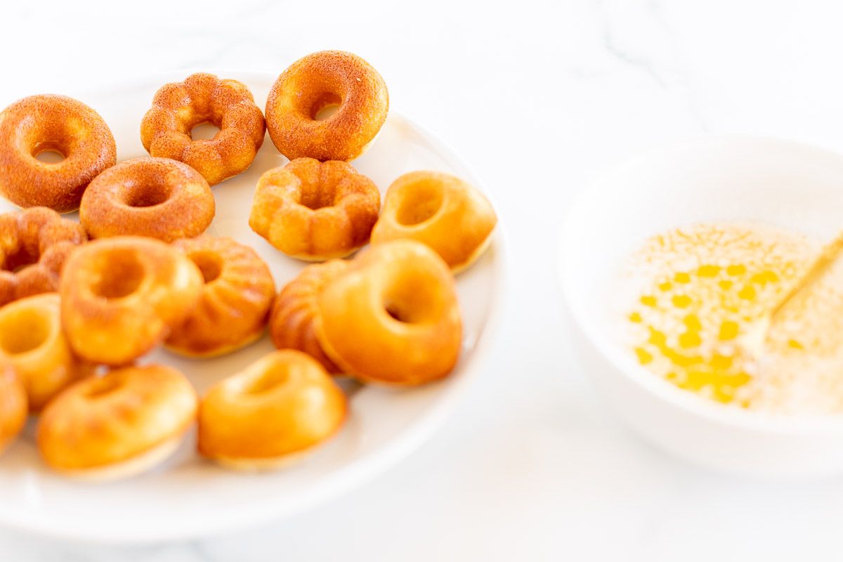 Baked cinnamon sugar donuts on a white platter, with a bowl of melted butter nearby.