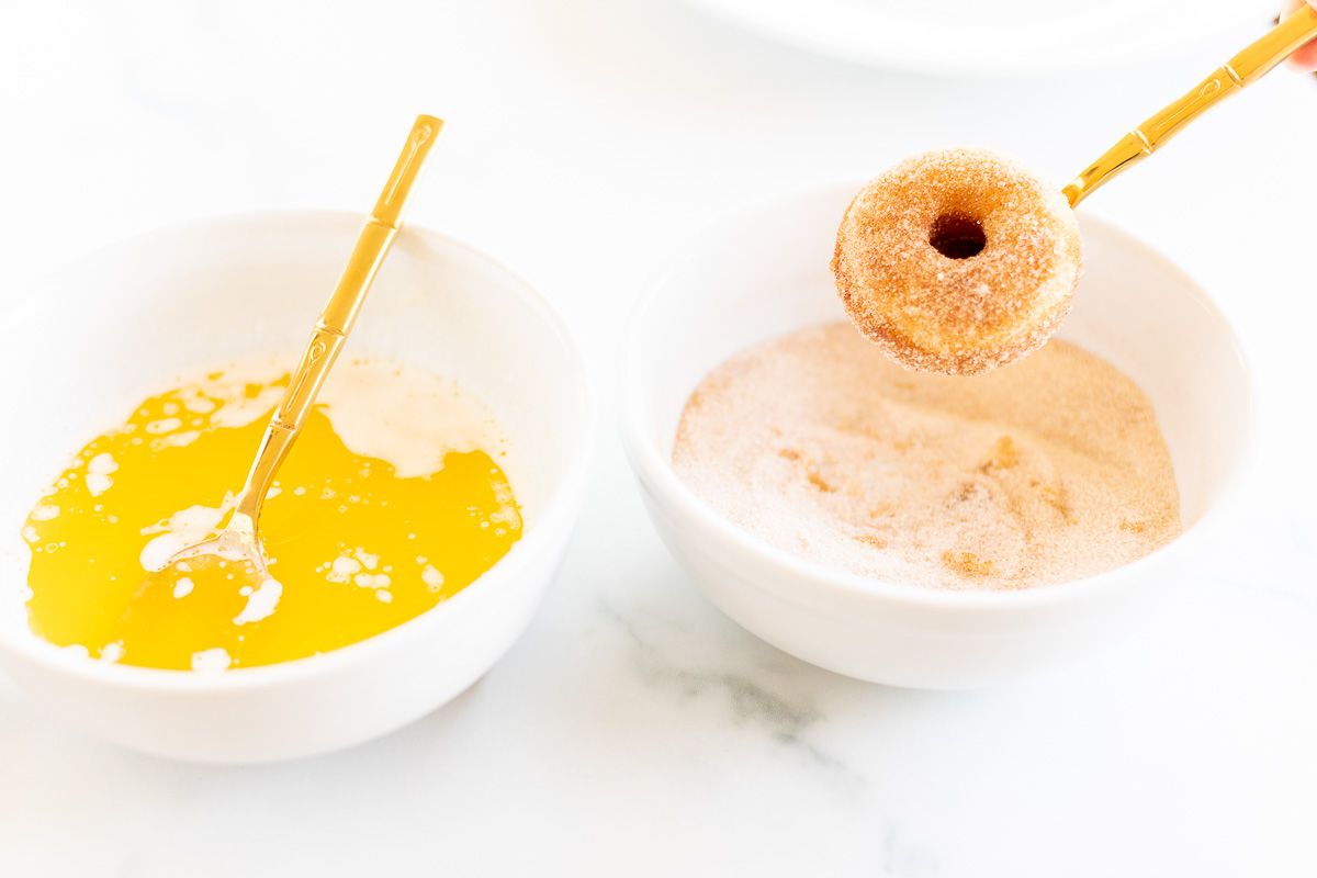 A baked donut being dipped into butter and a cinnamon sugar mixture.