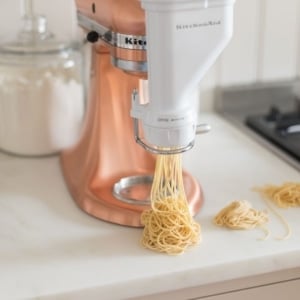A copper Kitchenaid mixer with a pasta attachment. A small pile of spaghetti shaped egg noodles are piling onto a marble countertop.