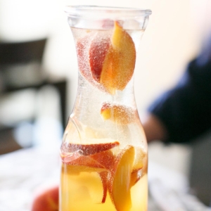 Prosecco Sangria in a glass carafe with pomegranate and peaches in the back.