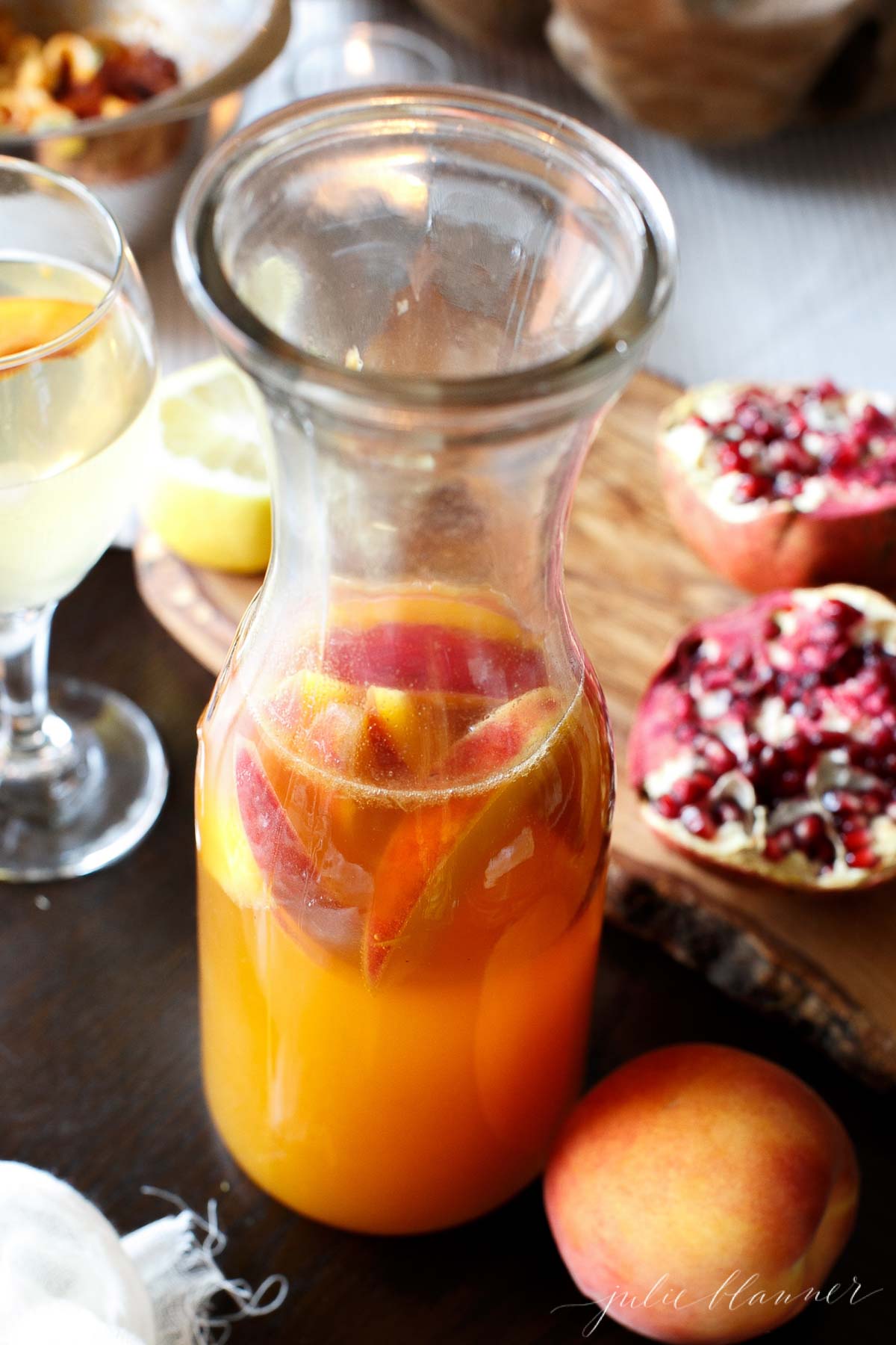 Prosecco Sangria in a glass carafe with pomegranate and peaches in the back.