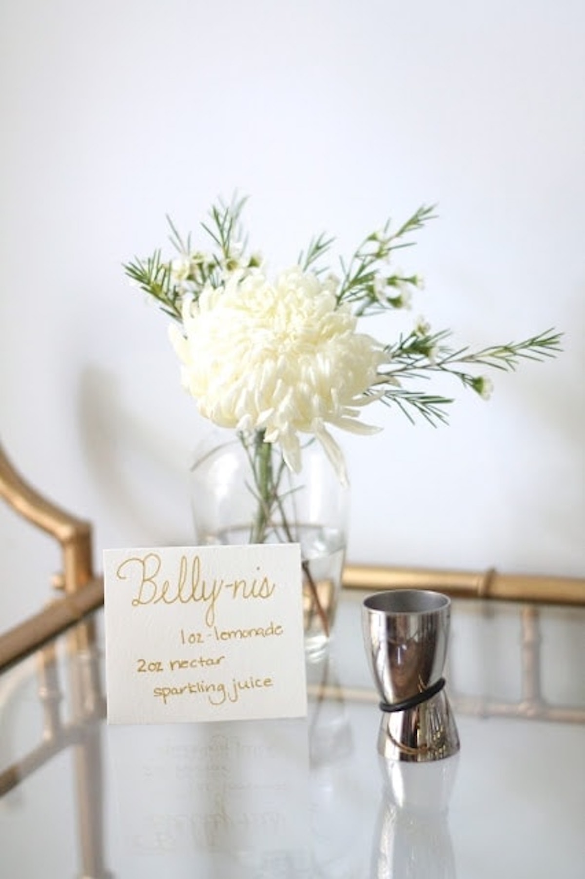 A flower arrangement with white flowers and green stems is in a glass vase on a reflective surface next to a stainless steel jigger and a recipe card for a Non-Alcoholic Bellini, perfect for a baby shower mocktail.
