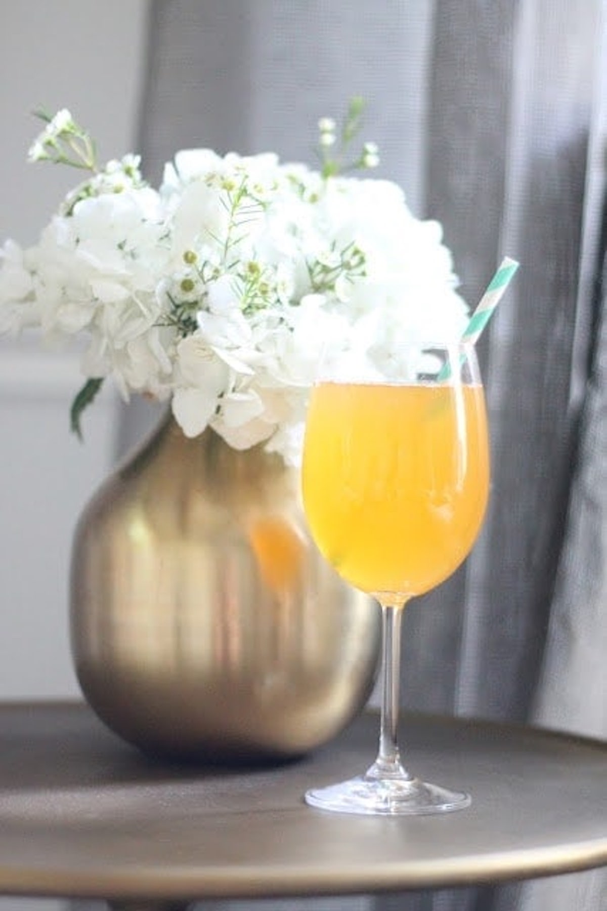 A yellow Non-Alcoholic Bellini with a green-and-white straw in a wine glass sits beside a golden vase of white flowers on a brown table, making it an elegant choice for any baby shower mocktail.