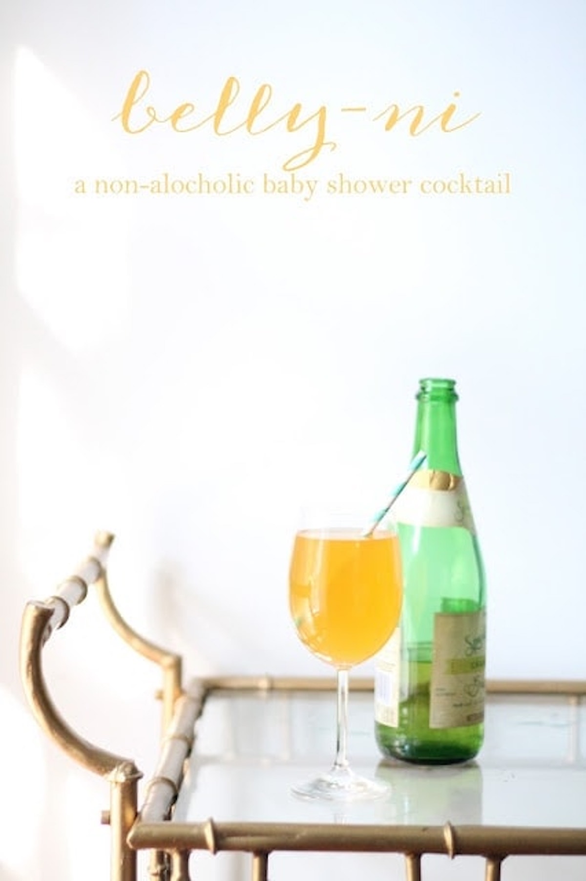 A non-alcoholic baby shower cocktail, cleverly named "belly-ni," is presented in a glass beside a green bottle on a gold-colored tray table.