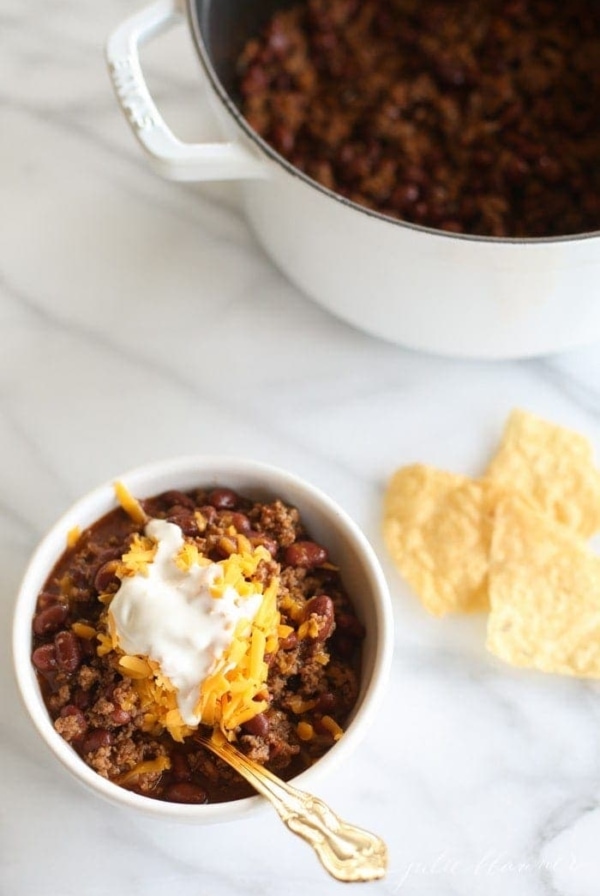A bowl of chili topped with cheese and sour cream, chips and the pot in the background.