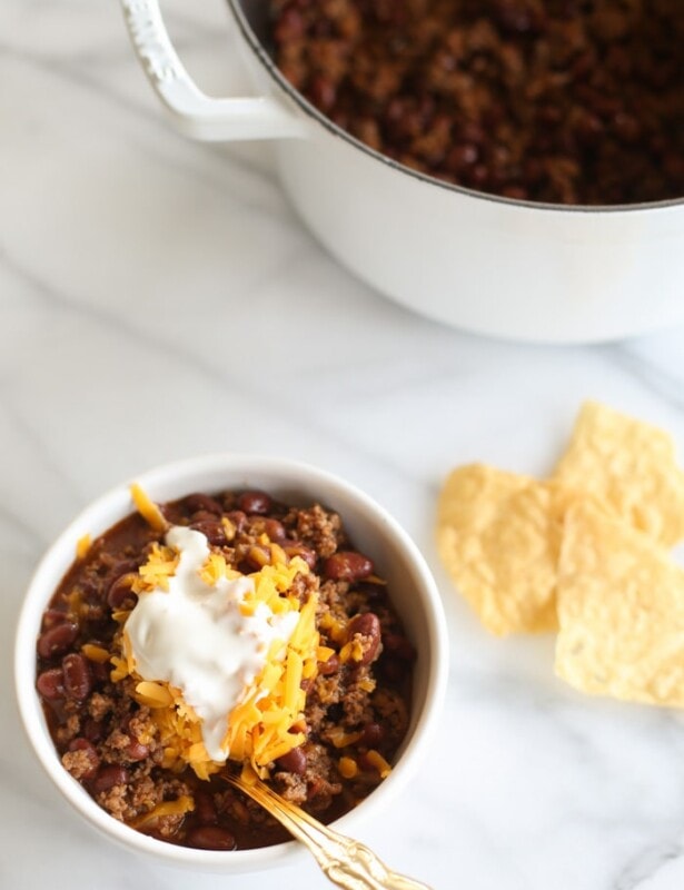A bowl of chili topped with cheese and sour cream, chips and the pot in the background.
