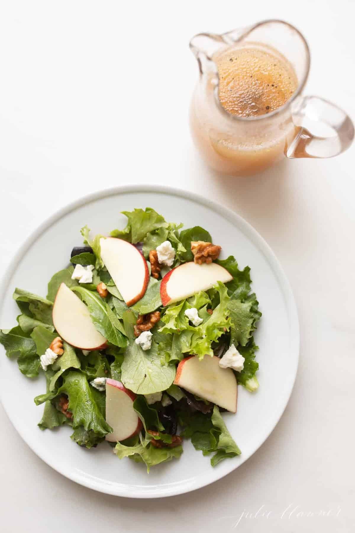 Apple cider vinegar dressing in a glass pitcher next to a plate of fresh salad. 