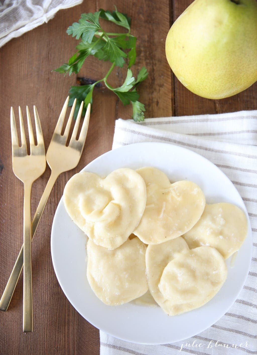 heart shaped ravioli on a white plate, gold forks to the side and a pear in the background.