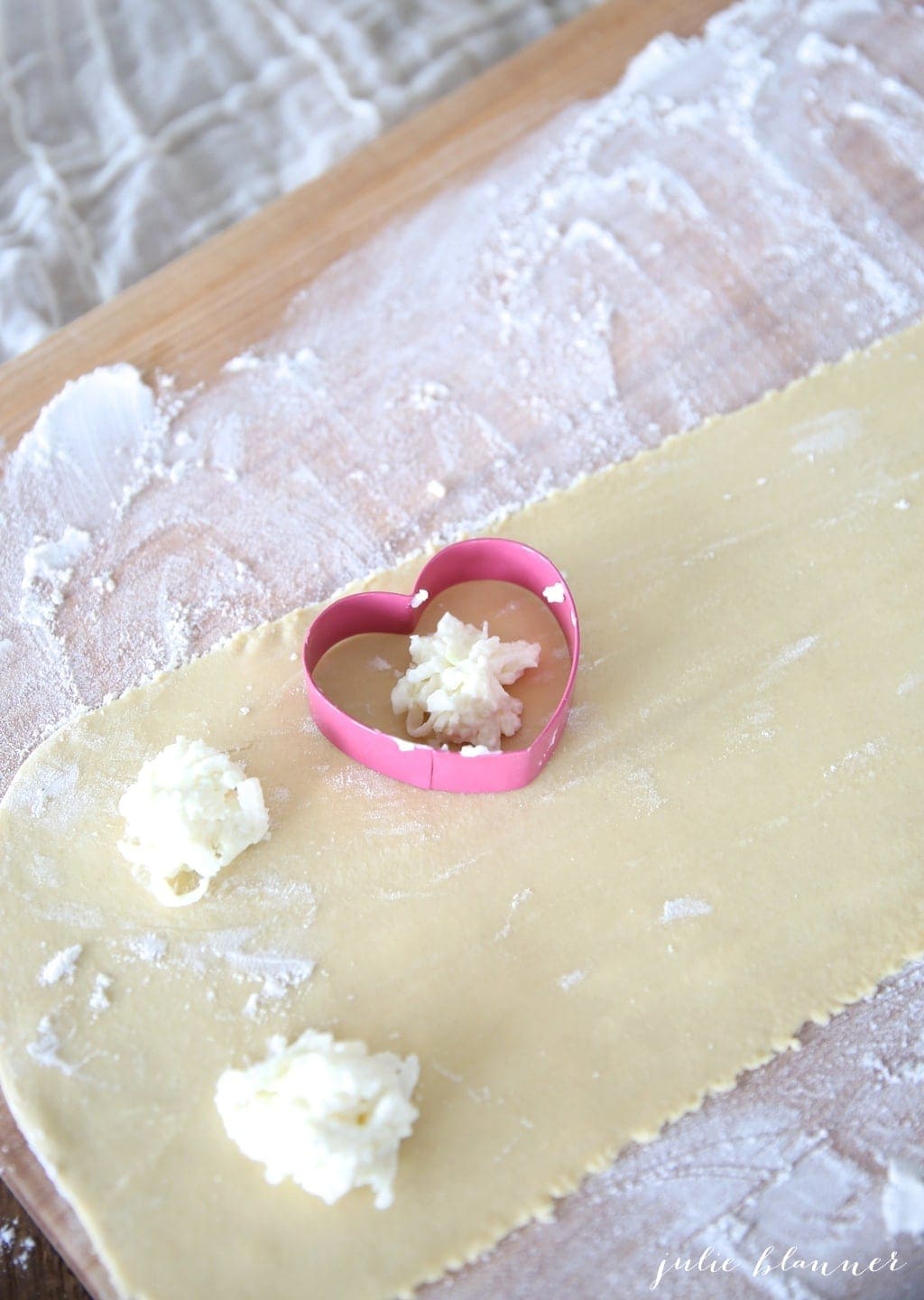 A thin sheet of pasta on a floured wooden surface, pink heart cookie cutter forming homemade ravioli.