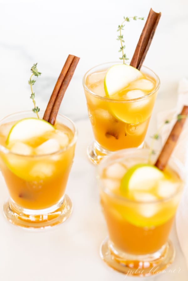 Three glasses filled with apple cider cocktails, garnished with apple slices and a cinnamon stick.
