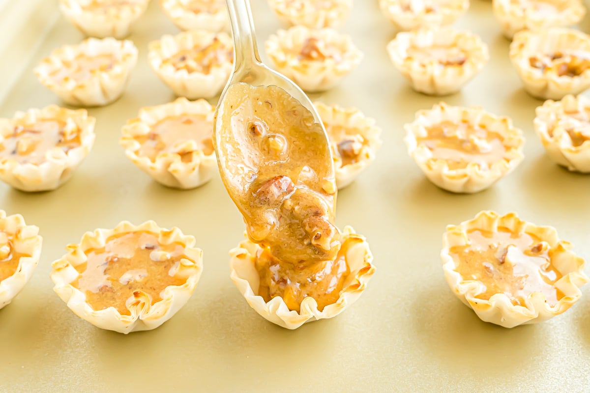 A spoon is drizzling caramel sauce on a tray of Mini Pecan Pie desserts.