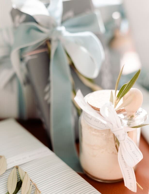 A clear glass jar of mint milk bath recipe under a tree with blue and wrapped gifts.