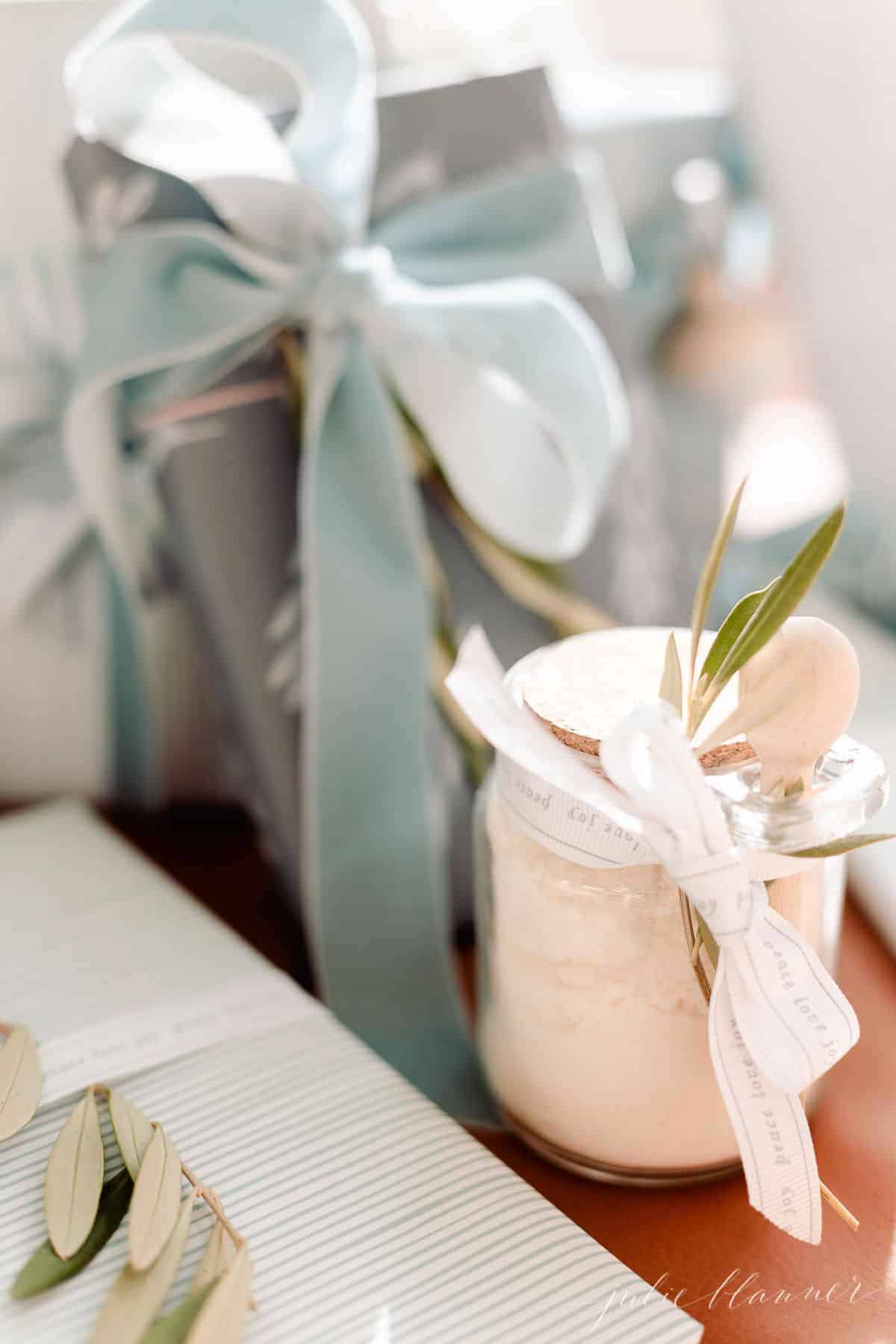 A clear glass jar of mint milk bath recipe under a tree with blue and wrapped gifts. 