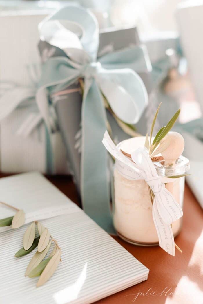 A clear glass jar of mint milk bath recipe under a tree with blue and wrapped gifts.
