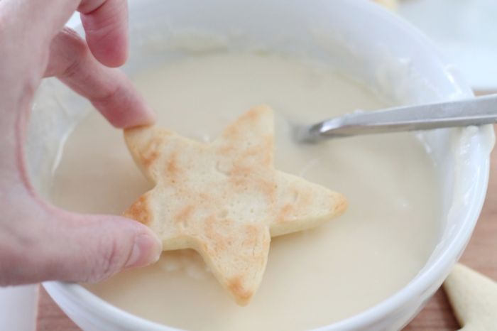 A hand dipping a star shaped cookie into a sugar cookie frosting recipe.