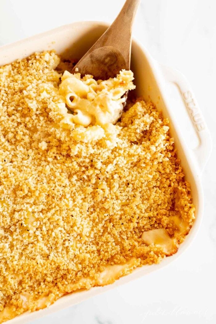 A white baking dish full of bacon mac and cheese, wooden spoon scooping at the edge.